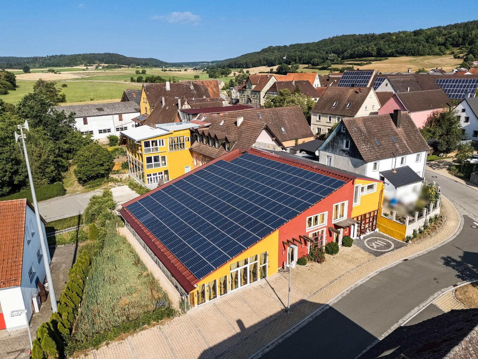 Luftaufnahme von Punkt XII im Steigerwald – das rot-gelbe Seminarhaus mit großer Solaranlage, eingebettet in Dorf, Felder und Wald